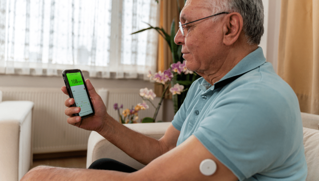 Ein älterer Mann, der zu Hause auf dem Sofa sitzt, hält ein Handy in der Hand. Auf dem Bildschirm wird der Wert seines Blutzuckers in einer App angezeigt.