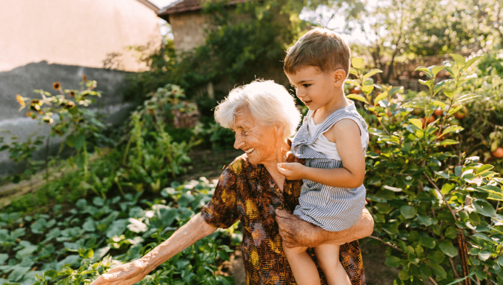 Eine 100-jährige alte Dame ist mit ihrem Urenkel im Garten und trägt ihn auf dem Arm.