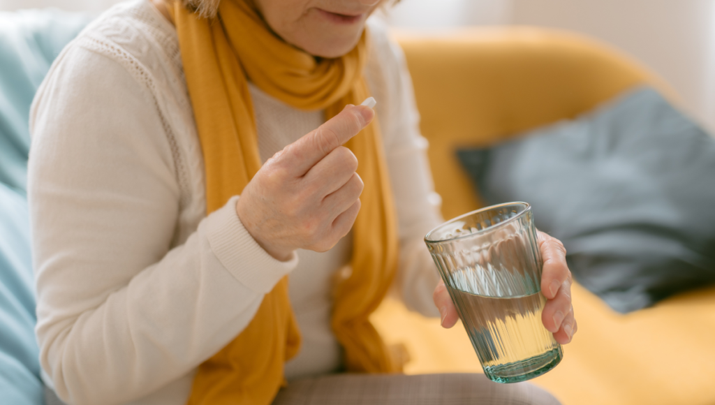 Antibiotika-Einnahme: Eine ältere Dame sitzt aufrecht und hält eine Tablette in der einen Hand, ein Glas Wasser in der anderen.