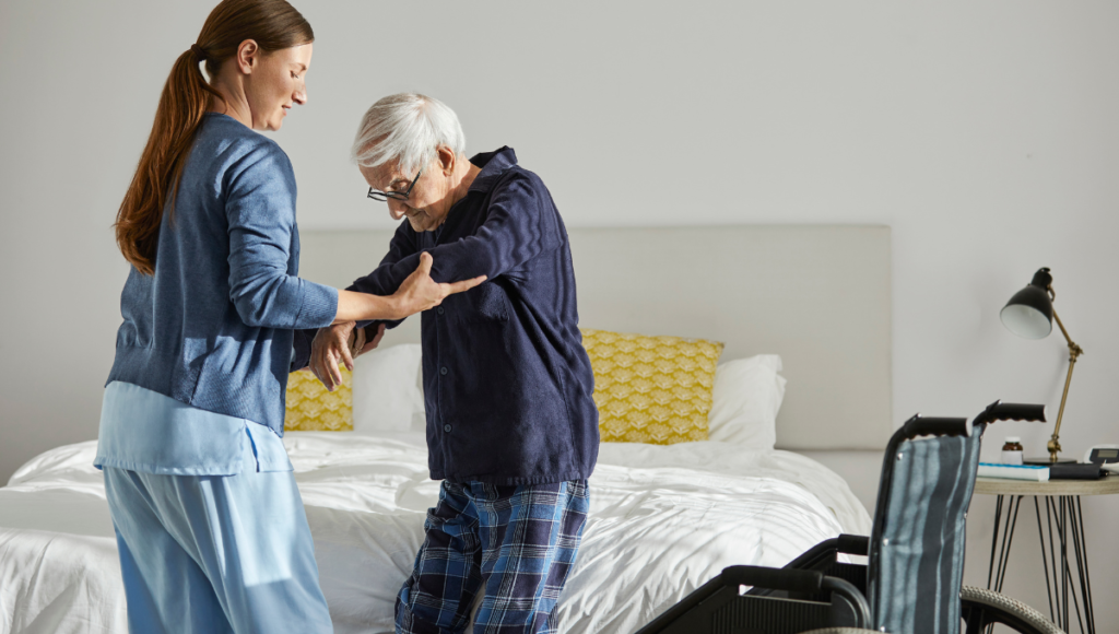 Eine Frau stützt einen älteren Mann und hilft ihm vom Bett in den Rollstuhl zu wechseln: Mit dem Konzept der Kinästhetik können Sie Mobilität und Selbstbestimmtheit Ihres pflegebedürftigen Angehörigen erhalten helfen.