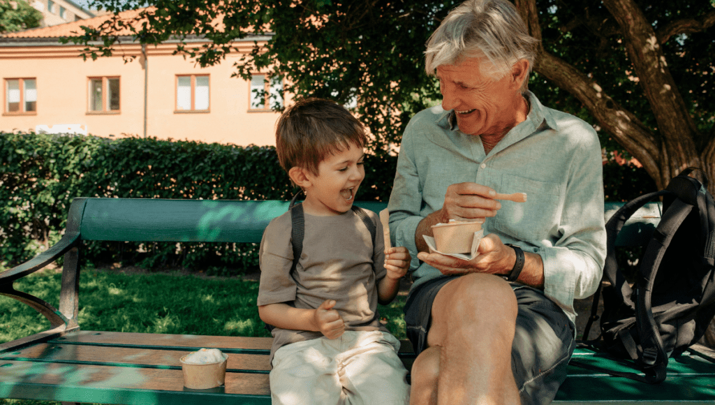 Ein Großvater sitzt mit seinem Enkel auf einer Bank und sie lachen gemeinsam, während sie Eis essen.