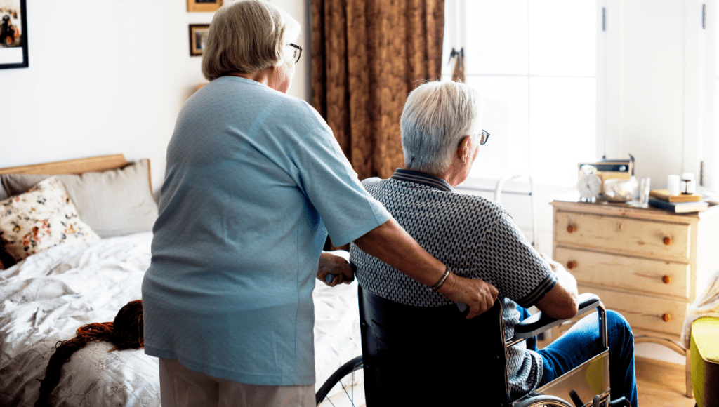 Eine Frau schiebt ihren Mann in einem Rollstuhl in das Pflegezimmer.